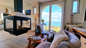 Ein gemütliches Wohnzimmer mit Kamin, bequemen Sitzmöglichkeiten, Meerblick durch große Fenster und rustikaler Einrichtung.