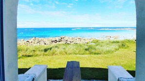 Eine hölzerne Bank mit Kissen, vor einem strahlend blauen Himmel gelegen, mit Blick auf einen felsigen Strand und eine Wiese.