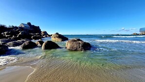 Ein Strand mit großen Felsen, klarem Wasser und einem Gebäude links.