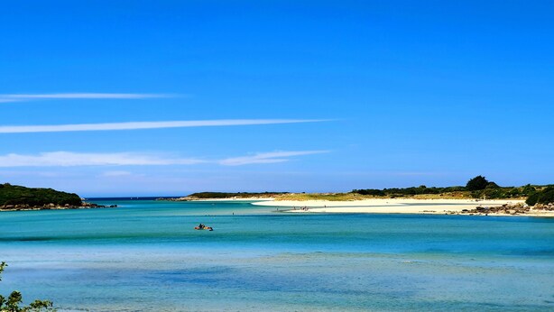 Een strand met helder turquoise water, een zandstrand en heuvels in de verte.