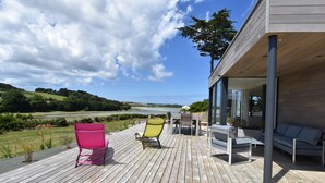 Een houten terras met buitenmeubilair, uitzicht op een rivier, en een modern gebouw met grote ramen.
