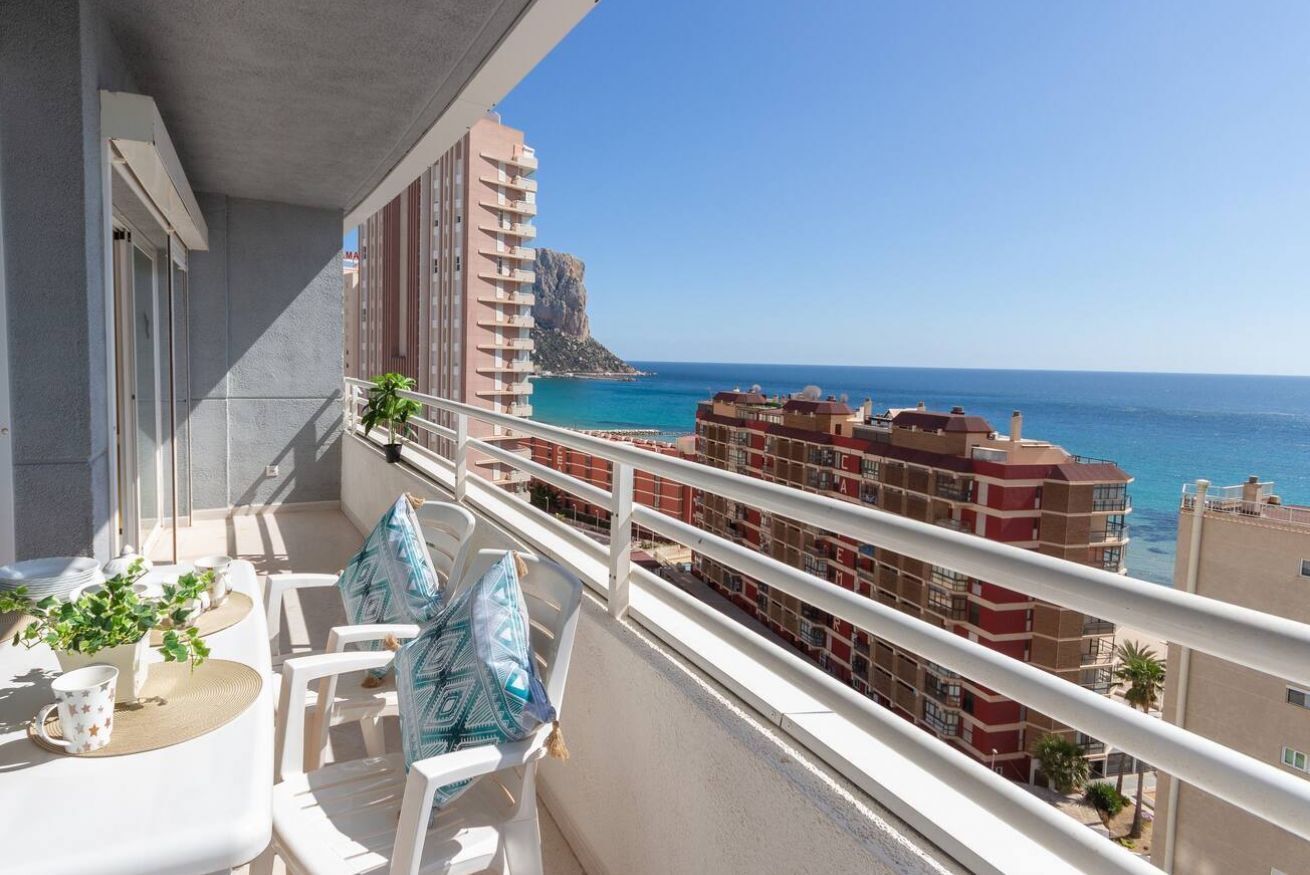 Een balkon met witte stoelen en een tafel, met uitzicht op de kust, gebouwen en een klif.