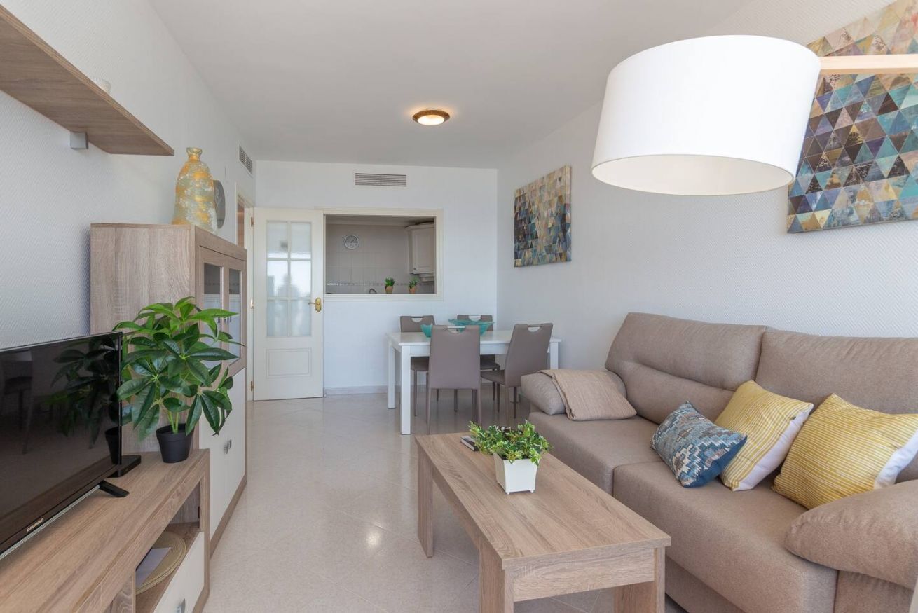 A modern living room with a flat-screen TV, a wooden coffee table, a beige sofa with patterned cushions, and a dining area in the background.