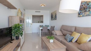 A modern living room with a flat-screen TV, a wooden coffee table, a beige sofa with patterned cushions, and a dining area in the background. - Apartamento Apolo XIV  by Costa CarpeDiem (Calp)