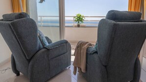 A room with two grey armchairs, a potted plant, and a view of the sea through large windows. - Apartamento Apolo XIV  by Costa CarpeDiem (Calp)