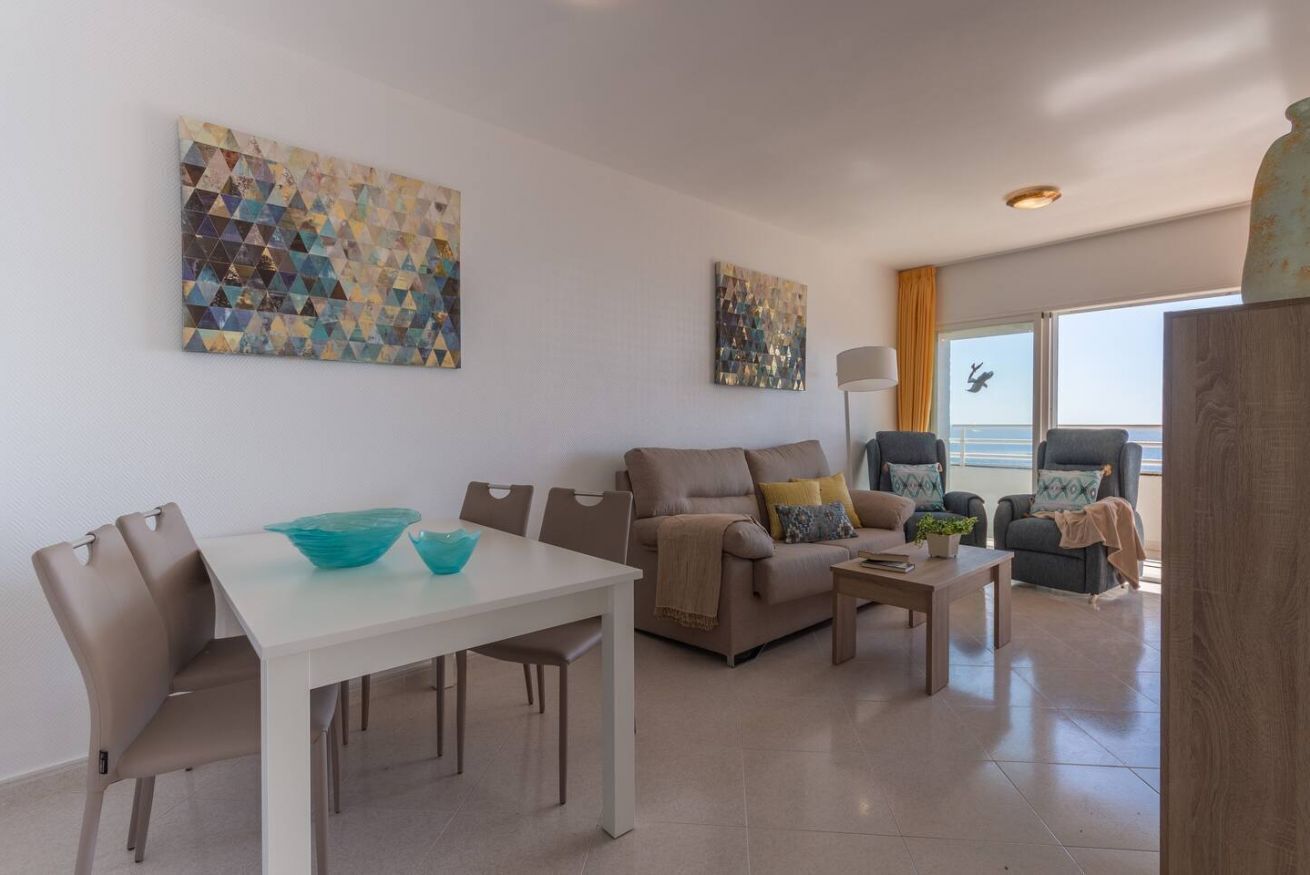 Una sala de estar moderna con una mesa de comedor blanca, sofá color beige y sillones. Hay cuadros abstractos en las paredes y un ventanal con vistas.