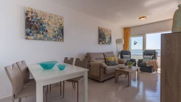 Una sala de estar moderna con una mesa de comedor blanca, sofá color beige y sillones. Hay cuadros abstractos en las paredes y un ventanal con vistas.