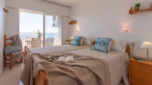 A bedroom with a large bed, a chair, a bedside table, and a view of the sea through a sliding door. - Apartamento Apolo XIV  by Costa CarpeDiem (Calp)