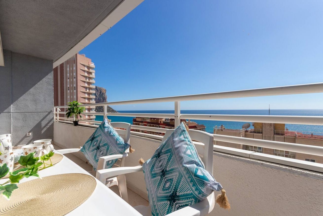 Een balkon met witte stoelen en een tafel, met uitzicht op een strand en de zee.