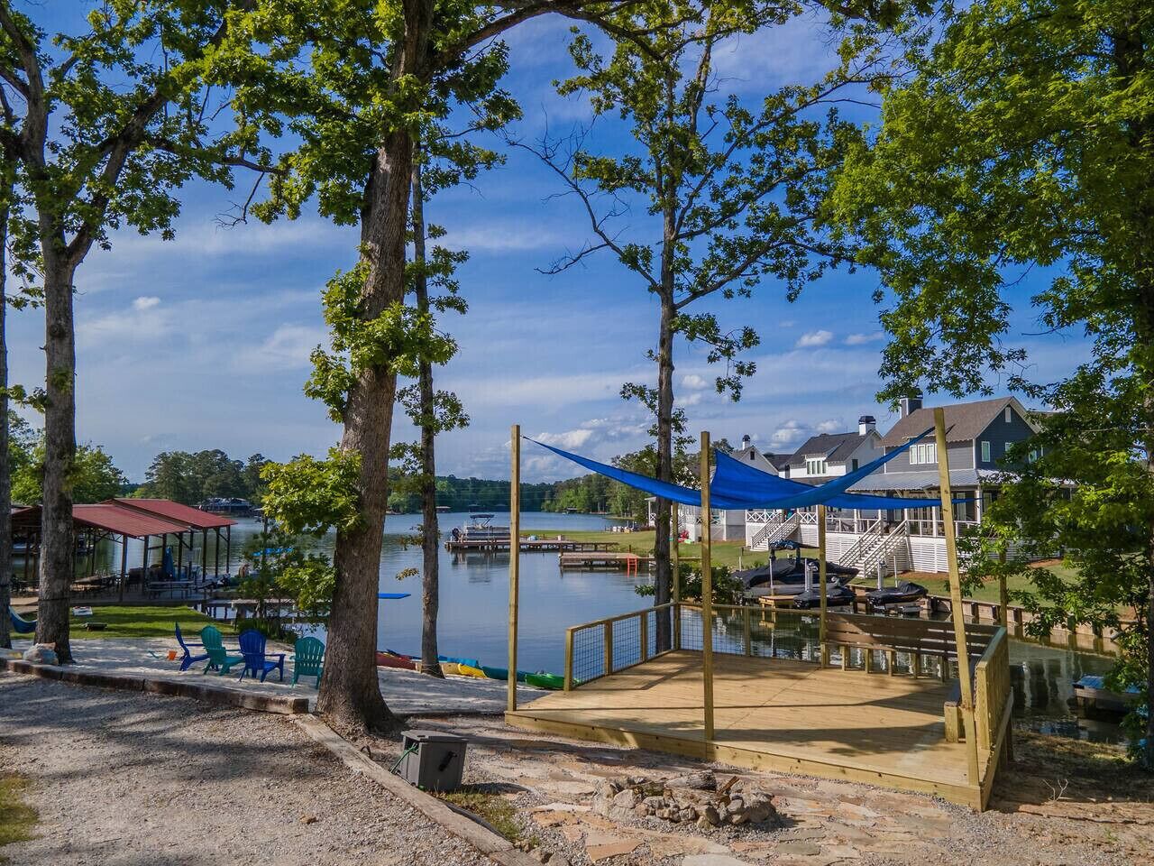 A lakeside park with a wooden deck, seating area, and a view of a house across the water.