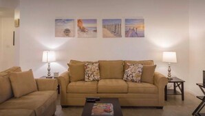 Un salon avec un canapé beige, deux tables d’appoint avec des lampes et trois œuvres d’art encadrées accrochées au mur.