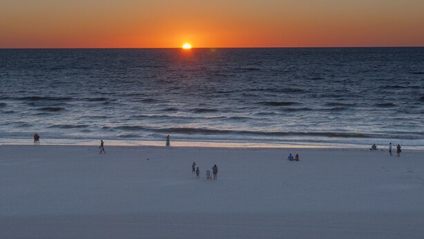 Une plage où le soleil se couche sur l’océan, avec des gens qui se promènent et qui sont assis sur le sable.