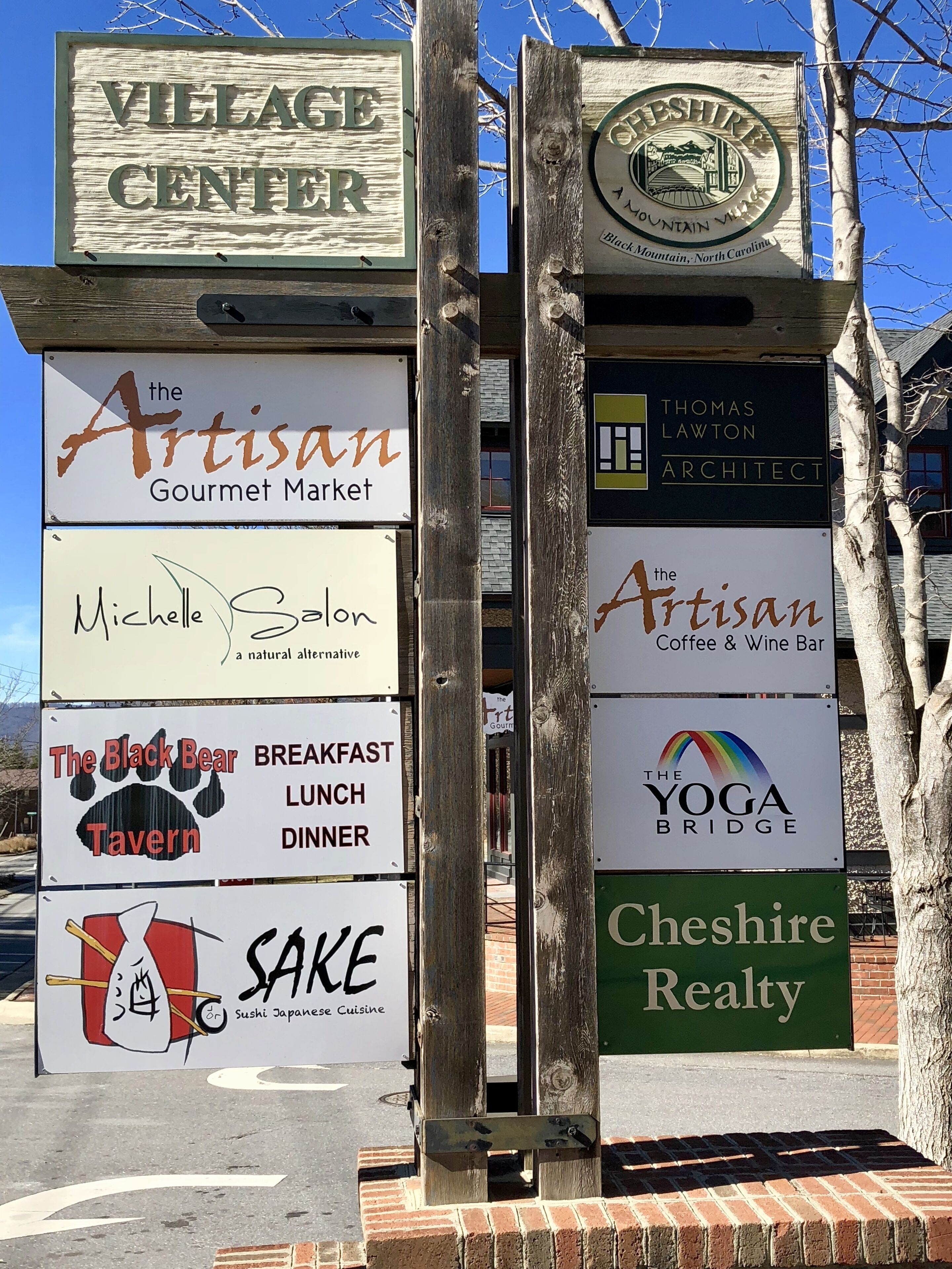 Un panneau indicateur portant divers noms d’entreprises, notamment « The Artisan Gourmet Market », « Michelle Salon », « The Black Bear Tavern », « Sake », « The Artisan Coffee & Wine Bar », « The Yoga Bridge » et « Cheshire Realty ».
