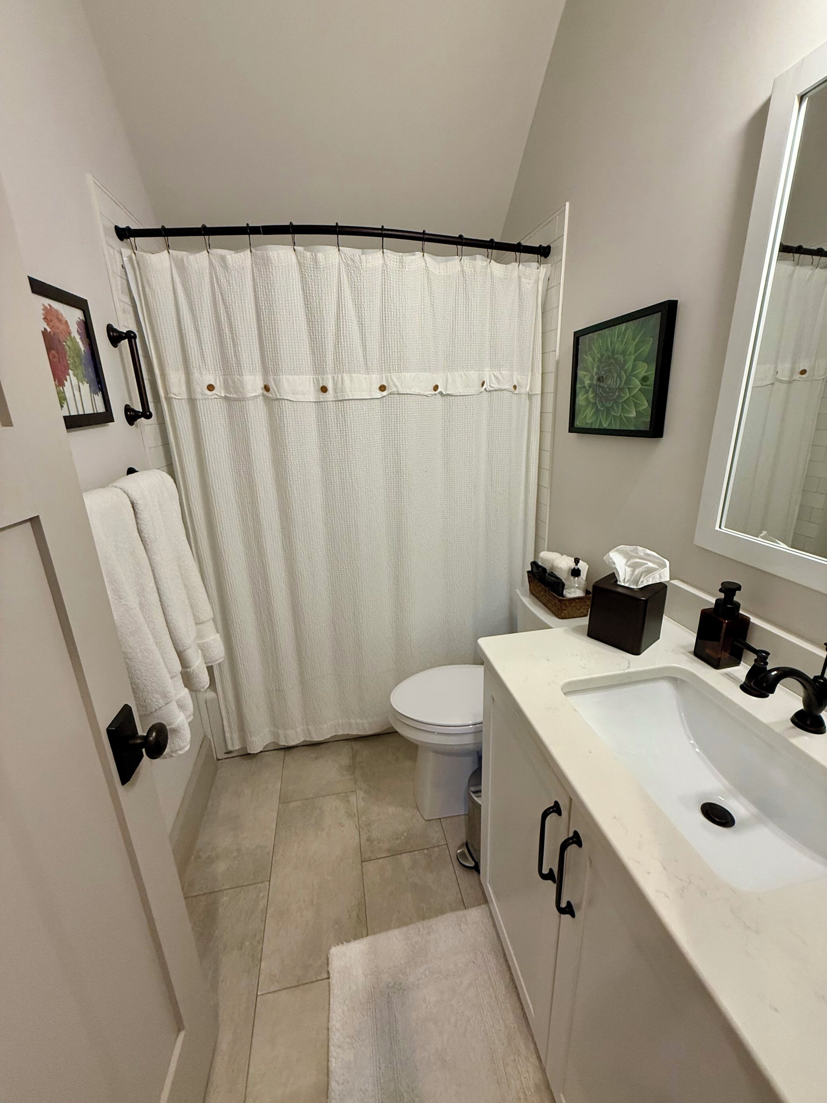 Un baño pequeño con lavamanos, inodoro y cortina de ducha blancos. Hay toallas colgadas en la pared y un cuadro enmarcado sobre el inodoro.