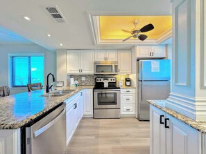 A modern kitchen with white cabinetry, stainless steel appliances, a granite countertop, and a ceiling fan. - Carlos Pointe #136 Tropical Beachfront Condo (Fort Myers Beach)