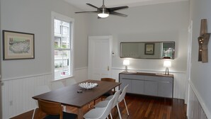 A dining area with a wooden table, white chairs, a ceiling fan, and a kitchen area with a mirror and two lamps. - Spacious home in the Historic District. Sleeps 8. Free Parking (Savannah)