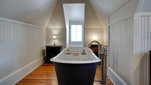 Una habitación estrecha con techos altos, una bañera grande, suelo de madera y una ventana con persianas.