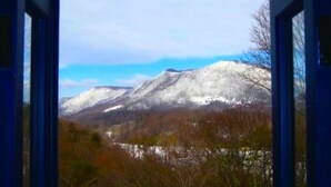 Vista desde una ventana con marco de madera, que muestra una cadena montañosa cubierta de nieve y un valle boscoso.