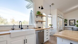 A modern kitchen with white cabinetry, a dishwasher, and a large window providing a view of the outdoors. - Avila Valley Luxury Estate with Pickleball Court (San Luis Obispo)