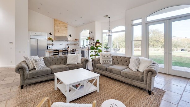 A spacious living area with a grey sectional sofa, a white coffee table, and a large window offering a view of the outdoors. - Avila Valley Luxury Estate with Pickleball Court (San Luis Obispo)