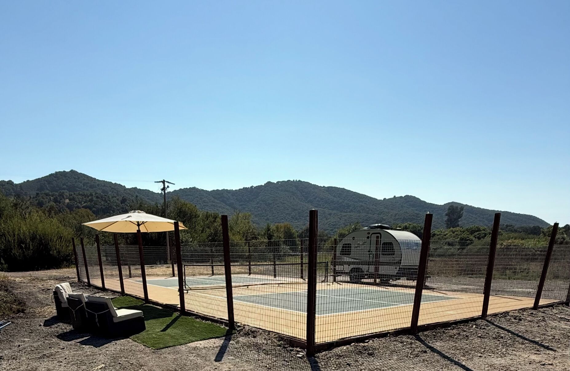 Ein eingezäunter Außenbereich mit Tennisplatz, überdachter Sitzgelegenheit und einem Wohnwagen.