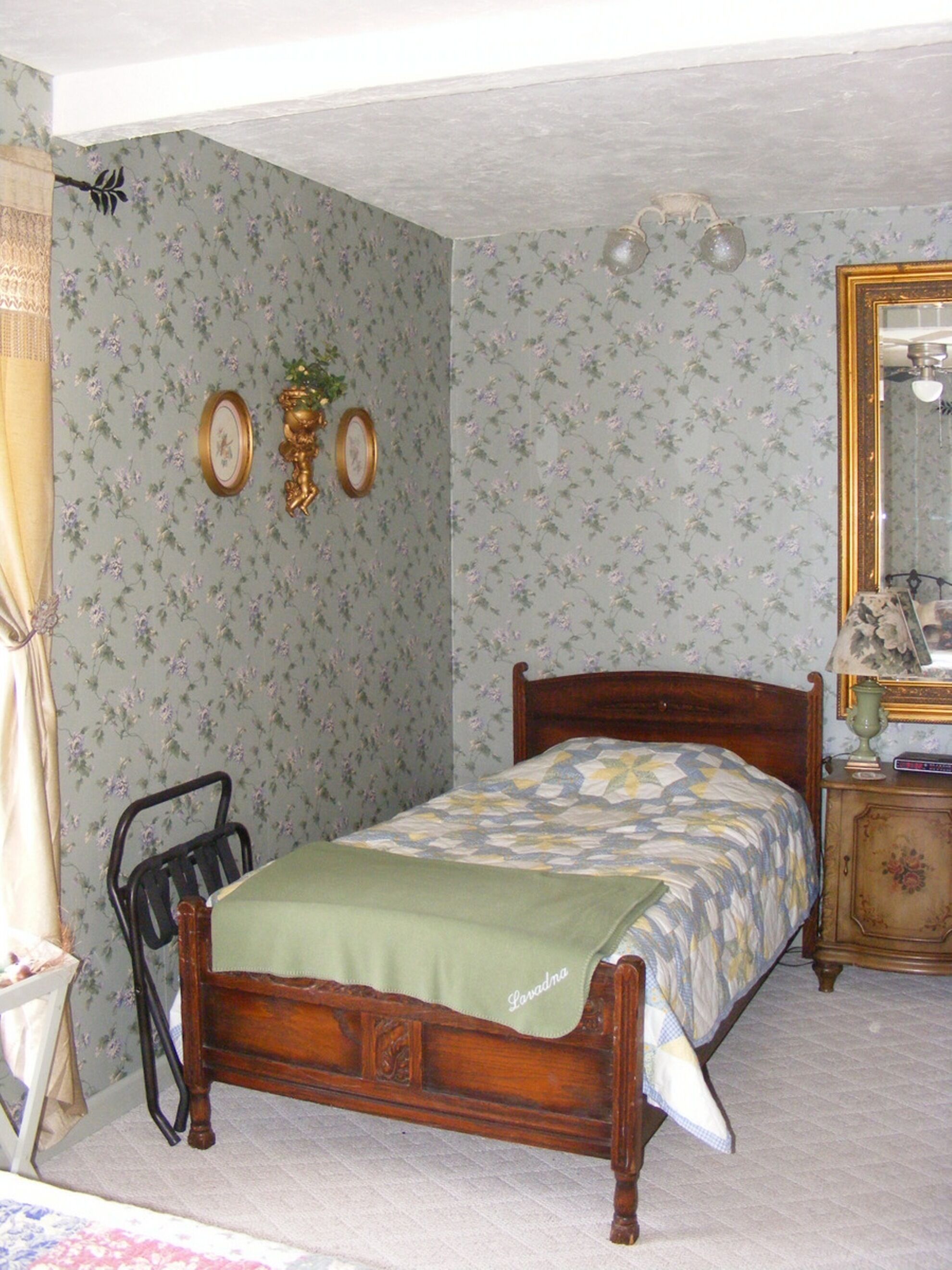 Une chambre avec un papier peint à motifs floraux, un lit en bois, une table de chevet avec une lampe et un miroir.