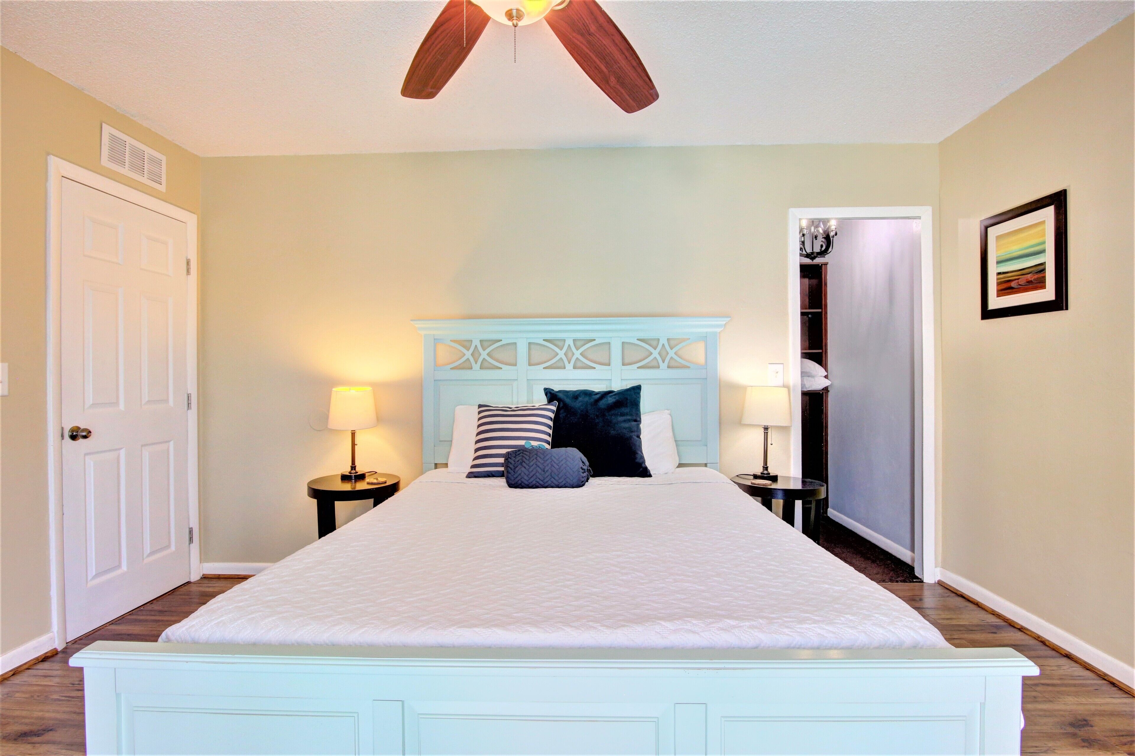 A bedroom with a bed, two bedside tables with lamps, a ceiling fan, a door, and a framed picture on the wall.