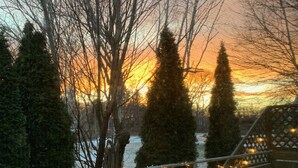Eine Holzterrasse mit Geländern und Lichterketten, mit Blick auf einen Fluss, an dessen Ufern sich kahle Bäume befinden, während die Sonne untergeht.