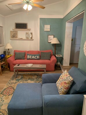 Un salon avec un canapé rouge, un canapé bleu et une table basse. On y trouve des éléments de décoration murale, une lampe et un ventilateur de plafond.