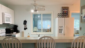 Een keuken met witte kastjes, een magnetron, een koffiezetapparaat en een koelkast. Er staan twee houten stoelen en een raam met jaloezieën.