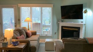 Een woonkamer met een open haard, een televisie erboven, een bank, een bijzettafeltje en een lamp.
