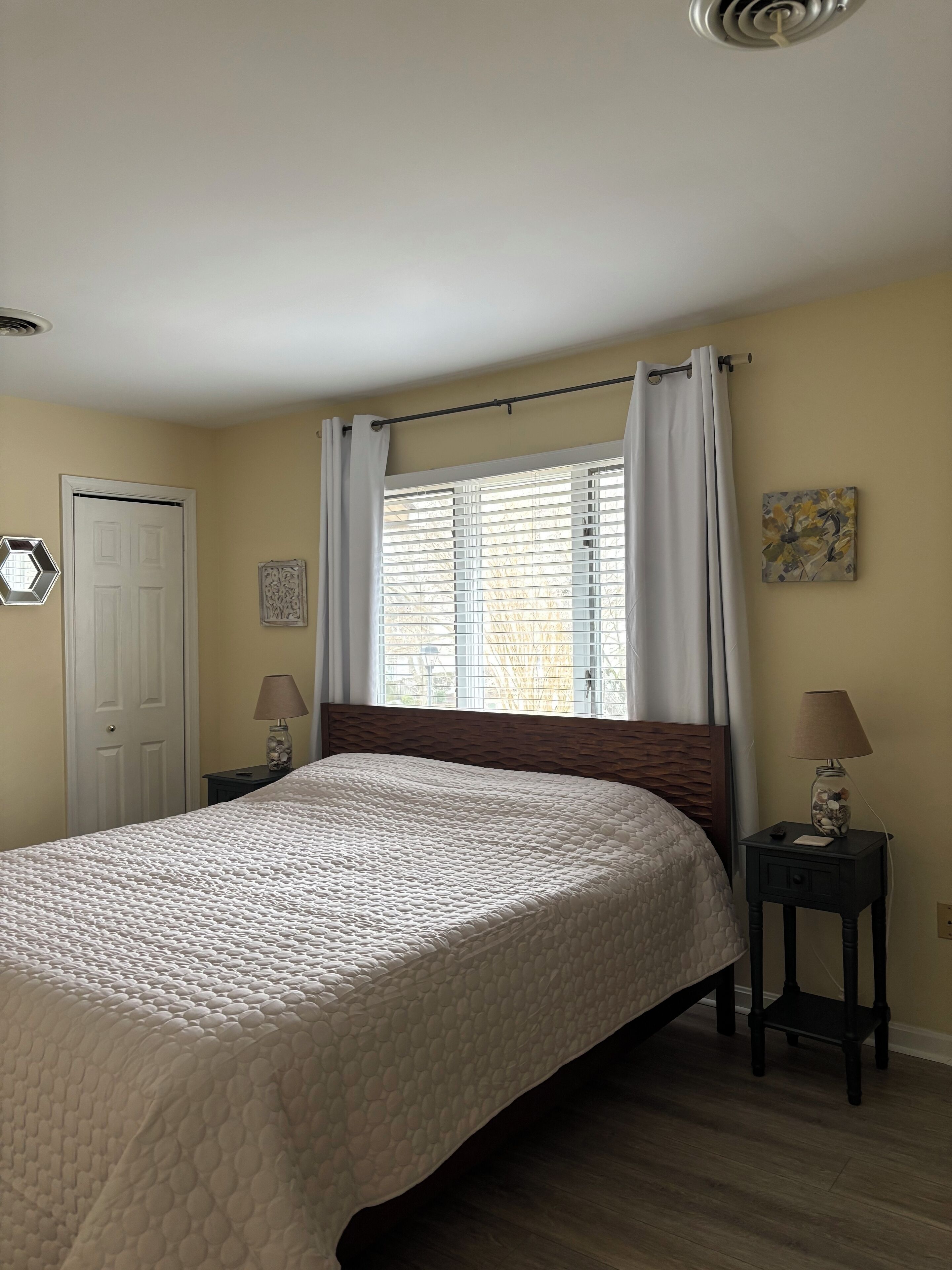 A bedroom with a bed, a window with curtains, a door, a nightstand with a lamp, and a wall decoration.