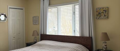 A bedroom with a bed, a window with curtains, a door, a nightstand with a lamp, and a wall decoration.