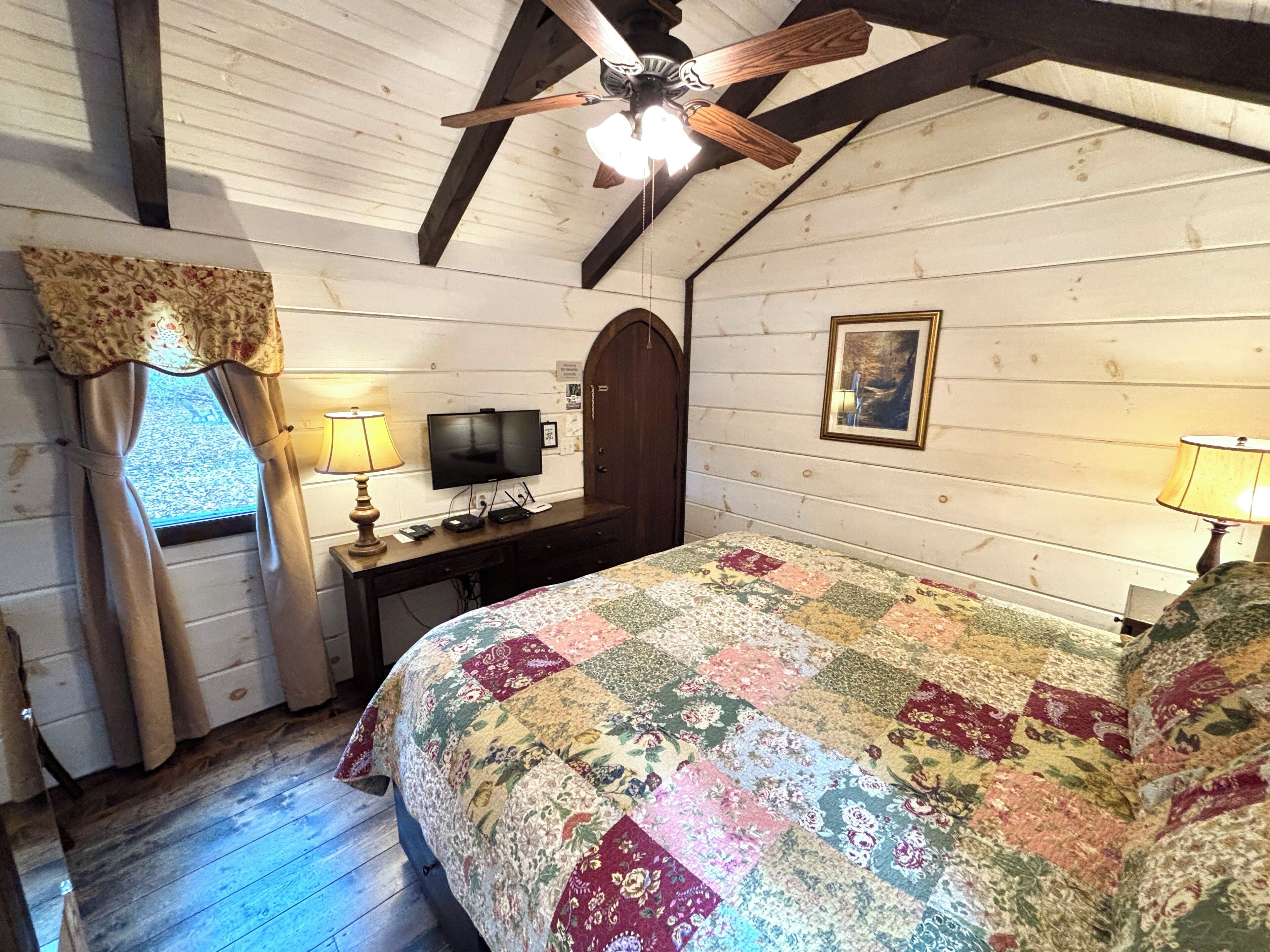 Une chambre avec un plafond en bois, un lit recouvert d’une couette aux couleurs vives, un bureau avec une télévision et une fenêtre avec des rideaux.