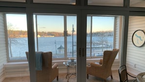 Une pièce avec vue sur un plan d’eau, deux fauteuils en osier, une petite table et une horloge au mur.