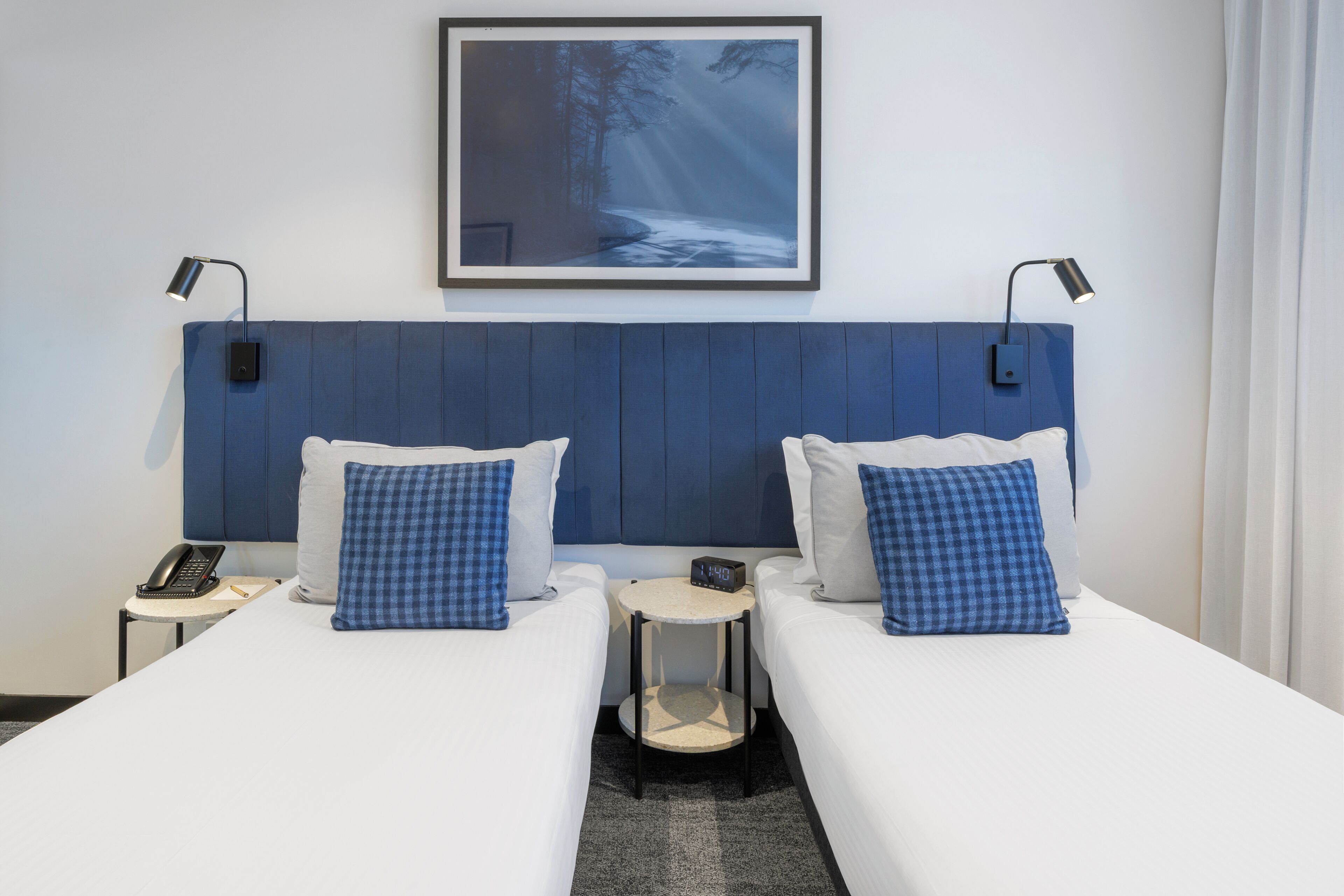 Une chambre d’hôtel avec deux lits, une table de chevet avec un téléphone, une horloge et un tableau encadré au mur.