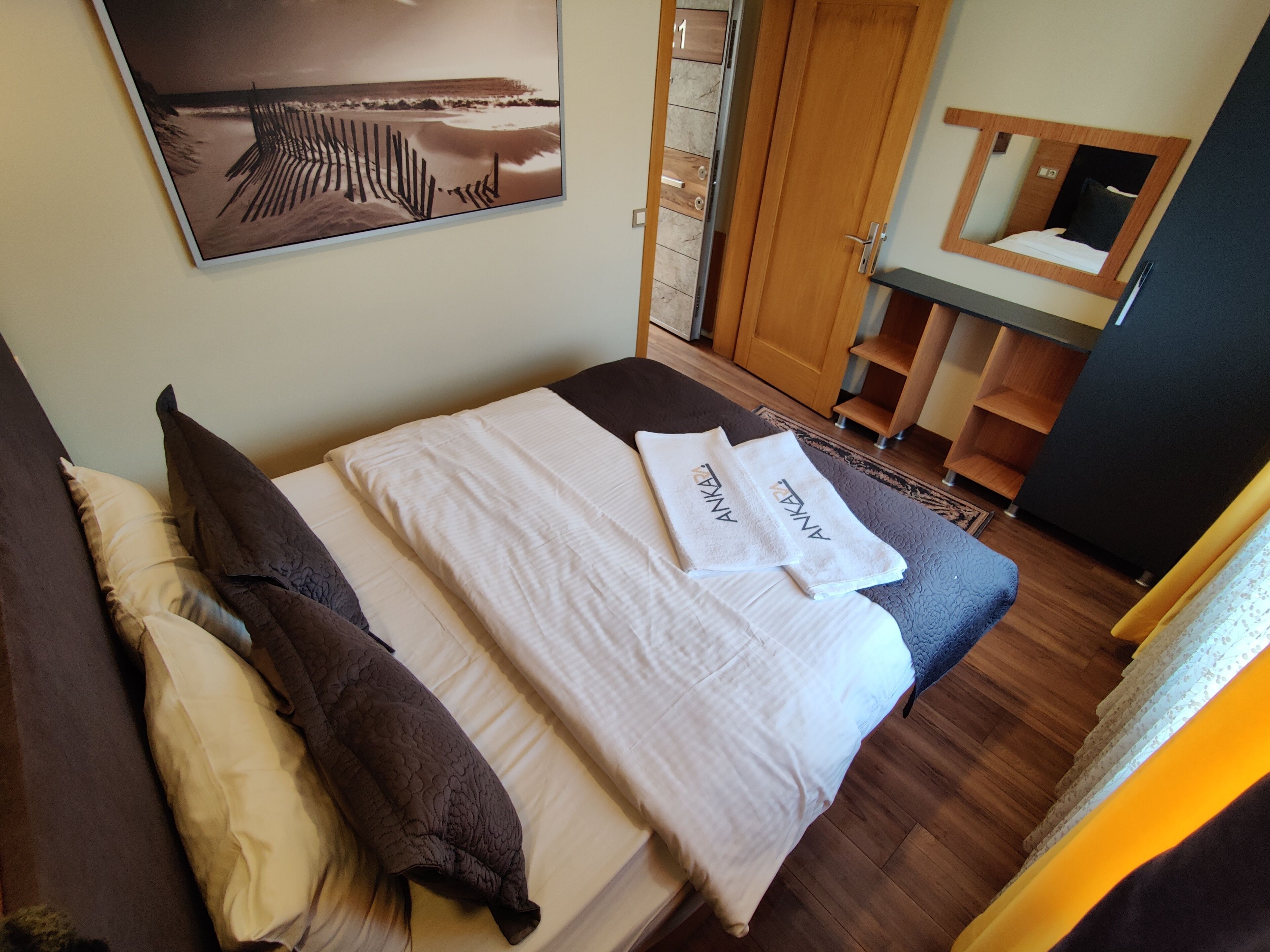 Een net opgemaakt bed met handdoeken en kussens, een houten garderobe, een kleine badkamer met spiegel en een wanddecoratie met een strandthema.