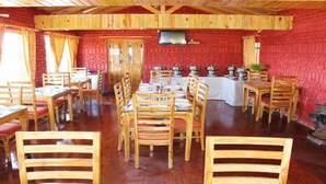 A dining area with wooden tables and chairs, a brick wall, and a television mounted on the wall. - Trinetra Orchards (Dhari)
