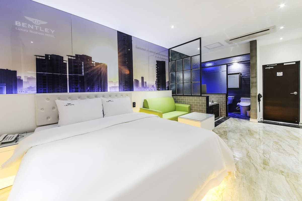 Ein Hotelzimmer mit einem großen Bett, einem grünen Sessel, einem kleinen Tisch, einem Stadtbild-Wandgemälde und einem durch eine Glasabtrennung einsehbaren Badezimmer.