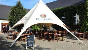 Un área de comedor al aire libre con la marca PAULANER, con mesas y sillas, un menú en una pizarra y un edificio al fondo.