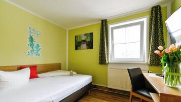 A hotel room with a bed, a desk, a chair, a window with curtains, a radiator, and a wall decoration.