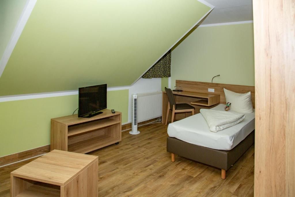 A small, single-room apartment with a sloped ceiling, a bed, a desk, a chair, a television, and a radiator.