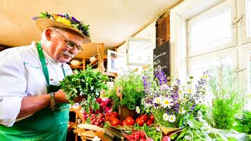 Una persona con delantal verde y sombrero de paja sosteniendo hierbas frescas en un puesto de mercado con diversas verduras y flores.