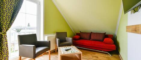 Una habitación con un sofá rojo, dos sillones, una pequeña mesa de madera y un ventanal con vistas.