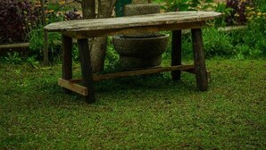 Un espacio de estar al aire libre rústico, con una mesa de madera y bancos, rodeado de follaje y una valla.