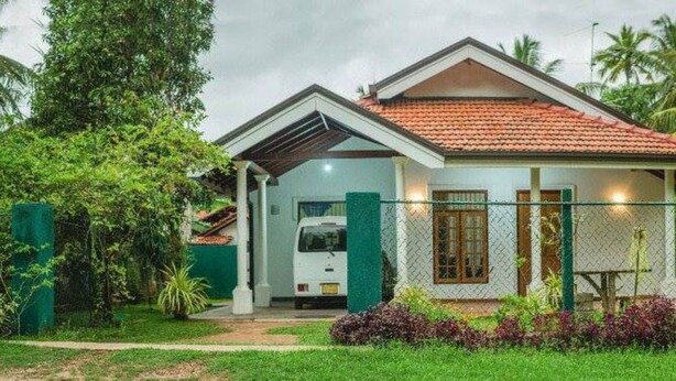 Una casa de un solo piso con techo de tejas rojas, rodeada de abundante follaje y palmeras.