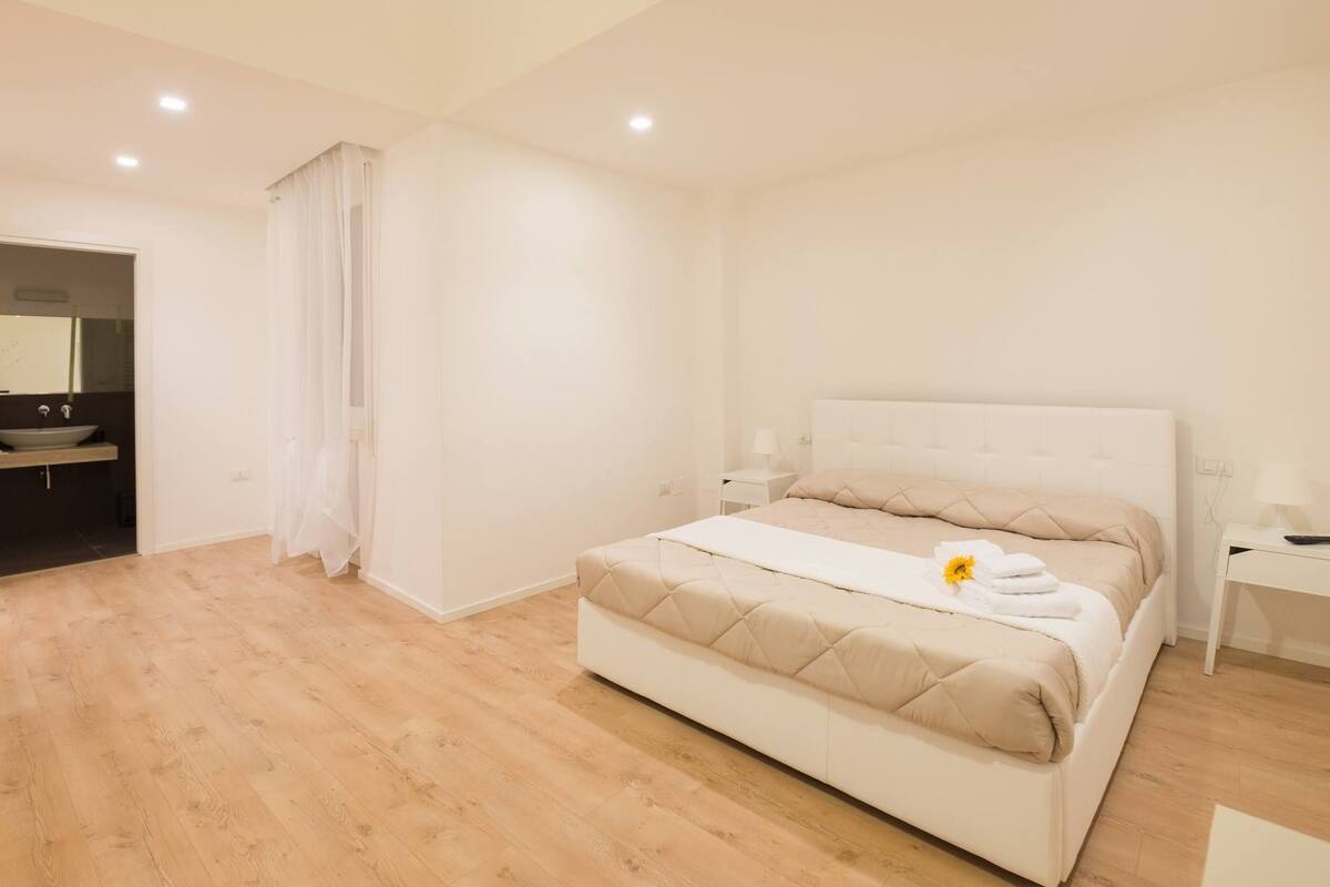 A modern bedroom with a large bed, a bedside table, a nightstand, and a bathroom visible through an open door.
