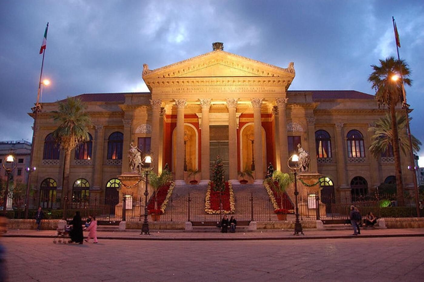 Un edificio grande de estilo clásico con una entrada central flanqueada por columnas, palmeras y una plaza bien iluminada.