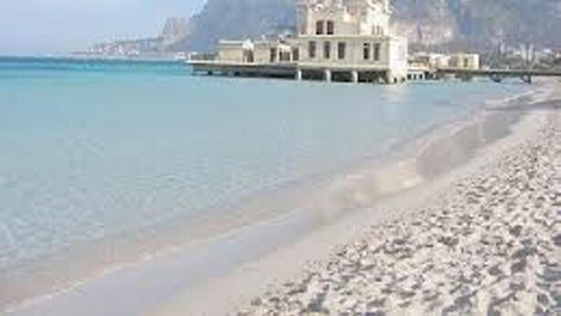 A beachfront property with a large building, clear blue waters, and a mountainous backdrop. - Dante 58 (Palermo)