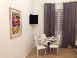 A dining area with a table set for four, white chairs, a framed colorful painting on the wall, a flat-screen TV mounted above the table, and a window with dark curtains.